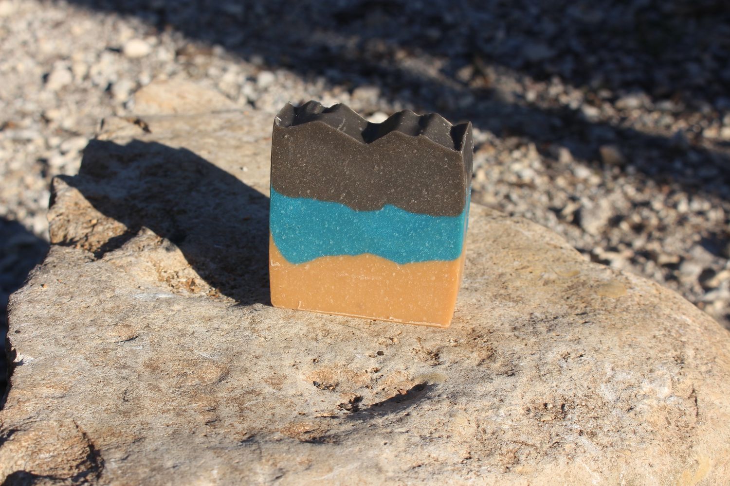 Handcrafted bar soap with three horizontal layers in tan, blue, and dark brown, standing upright on a stone surface.