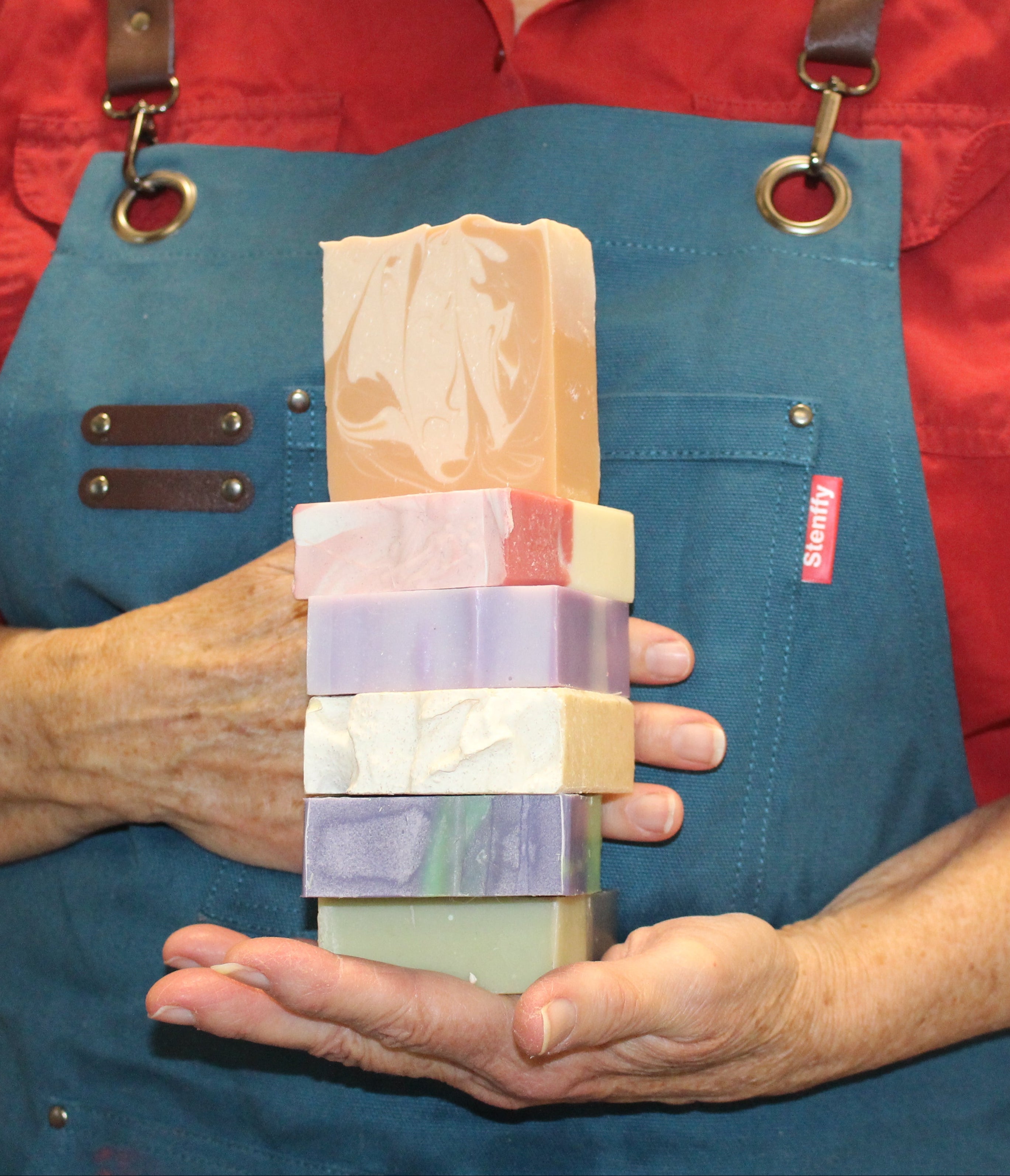 SS holding a stack of handmade soap bars wearing a red shirt and blue apron.